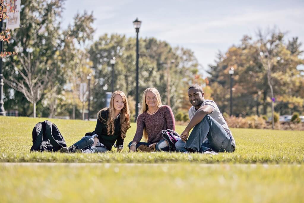 Three students sitting on the grass smiling at the camera